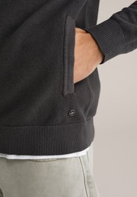 Temno sivi teksturirani pulover z rebrastim robom in manšetami. Ima stranski žep in majhen logotip na spodnji levi strani.