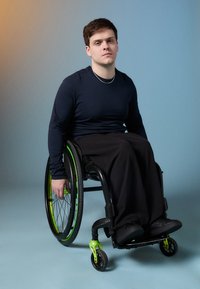 Egy fekete keretes és élénk zöld részletekkel ellátott kerekesszékben ülő személy, sötét hosszúujjú pólót és fekete nadrágot visel, a háttérben egy kék fal áll.