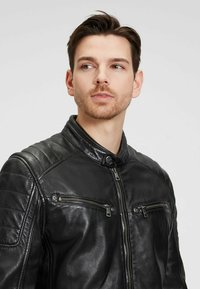 Zwarte leren motorjack met gewatteerde schouderdetails, staande kraag en ritszakken. Glad textuur en slim fit ontwerp.