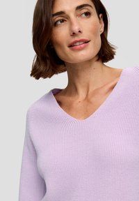 Lichtpaarse gebreide trui met een diepe V-halslijn en ribbeltextuur, met lange mouwen. Te zien op een model met kort haar.