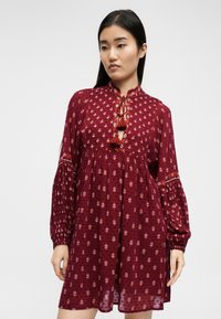 Femme portant une robe longue à manches longues de couleur bordeaux avec de petits imprimés floraux et des liens à pompons au niveau de l'encolure, sur un fond uni.