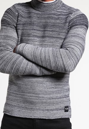 Homme les bras croisés portant un pull en maille côtelée rayé gris et noir et un pantalon noir sur un fond uni.