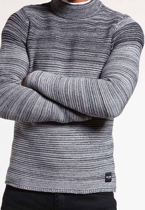 Homme les bras croisés portant un pull en maille côtelée rayé gris et noir et un pantalon noir sur un fond uni.