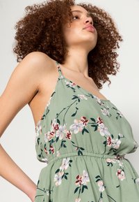 Vestido floral verde com alças finas, cintura elástica, apresentando flores rosa e brancas num tecido texturizado.