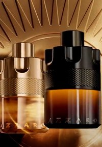 Deux flacons de parfum Azzaro, un doré et un noir, avec des bouchons texturés sur un fond doré rayonnant.