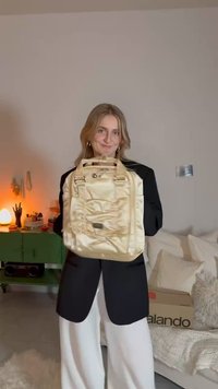 Gold satin backpack with multiple pockets, square shape, dual top handles, and a decorative charm. Worn by a person in a black blazer.