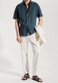 Lyhythihainen tummansinisenvihreä napitettava paita yhdistettynä kermaan vivahtaviin housuihin ja ruskeisiin sandaaleihin, kädessä vaaleanvärinen takki. Sileä kangaspinta.