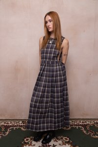 Frau mit langen roten Haaren, die ein ärmelloses lila kariertes Maxikleid und schwarze Schuhe trägt, steht vor einer beigen Wand auf einem gemusterten Teppich.