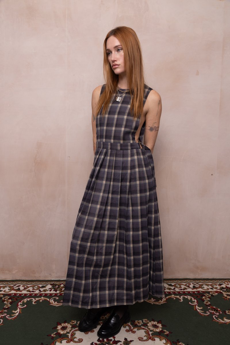 Frau mit langen roten Haaren, die ein ärmelloses lila kariertes Maxikleid und schwarze Schuhe trägt, steht vor einer beigen Wand auf einem gemusterten Teppich.