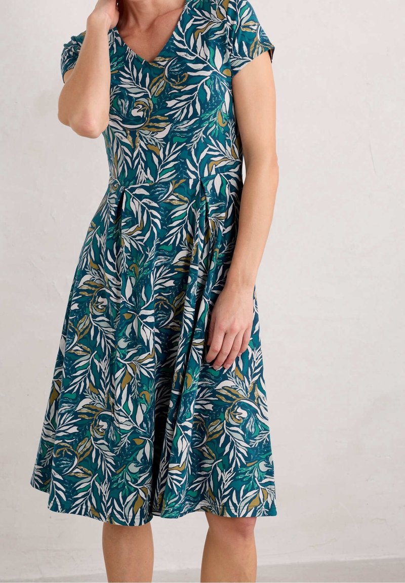 Femme portant une robe courte aux manches courtes, longueur genou, avec un motif feuillu de couleur sarcelle, blanc et or, debout contre un mur clair uni.