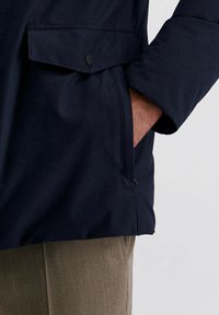 Marineblauwe jas van een gestructureerde stof, met een borstzak met klep en knop, gedragen over lichtbruine broeken.