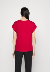 Top rosso a maniche corte con scollatura rotonda, texture morbida e vestibilità comoda. Indossato con pantaloni neri, visto da dietro.
