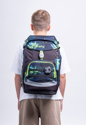 Kind trägt einen schwarzen und blauen Rucksack mit grünen Akzenten und fußballbezogenem Schlüsselanhänger, steht vor einem schlichten weißen Hintergrund, mit dem Rücken zur Kamera.