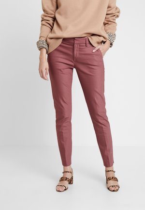 Person trägt mauve-farbene, eng anliegende Hose, beigen Pullover mit leopardenmusterartigen Bündchen und Sandalen mit Leopardenmuster und Blockabsatz, steht vor weißem Hintergrund.