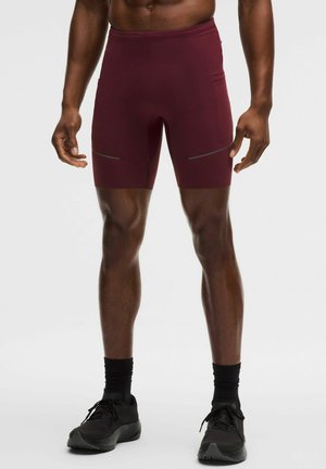 Atleta maschio che indossa pantaloncini a compressione color amaranto con tasche laterali e scarpe da ginnastica nere, in piedi davanti a uno sfondo neutro.