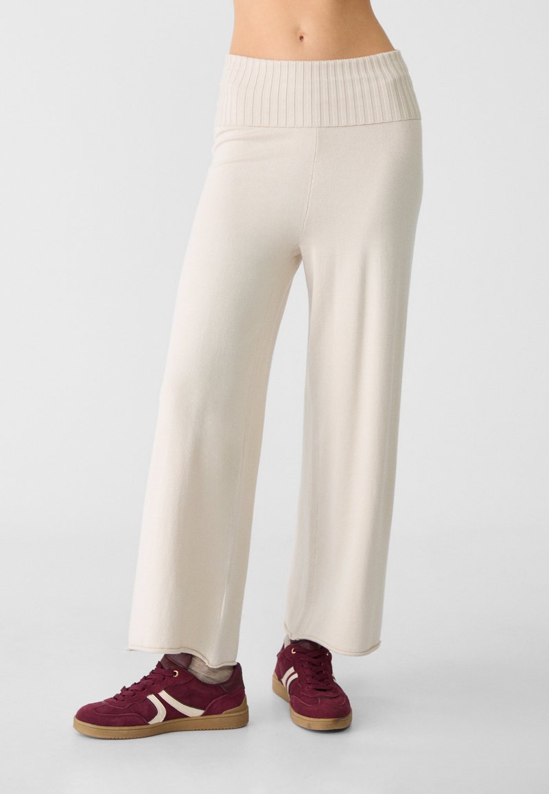 Pantaloni beige a gamba larga con una vita alta a costine, caratterizzati da una texture di tessuto morbido. Abbinati a sneakers bordeaux su uno sfondo di pavimento liscio.