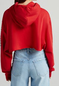 Sudadera corta roja con capucha ajustable, que cuenta con mangas largas y un dobladillo sin terminar, combinada con unos jeans de denim azul claro de cintura alta.
