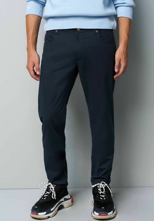 Marineblauwe slim-fit broek van rekbaar materiaal, met voorzakken en een knoopsluiting, gecombineerd met zwarte sneakers en witte veters.