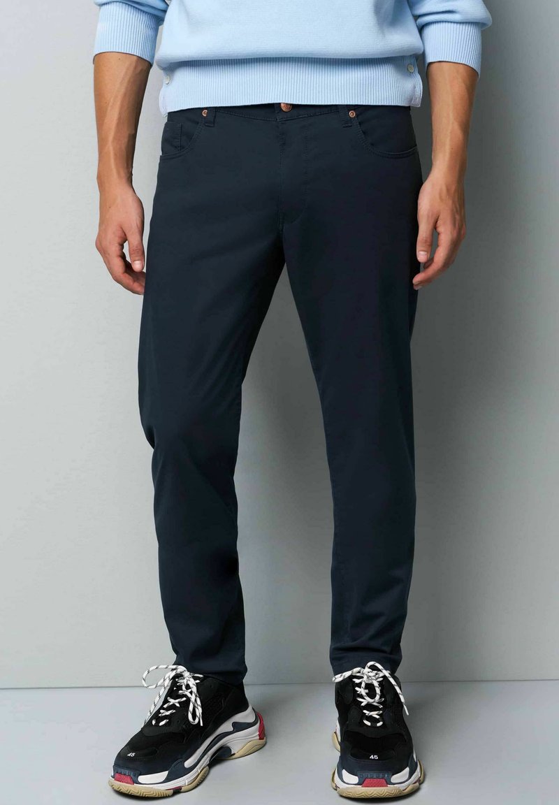 Marineblauwe slim-fit broek van rekbaar materiaal, met voorzakken en een knoopsluiting, gecombineerd met zwarte sneakers en witte veters.