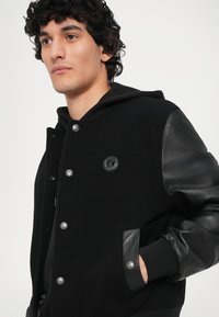 Jacheta varsity neagră cu corp din lână și mâneci din piele, cu nasturi tip snap, glugă și un logo circular pe piept.