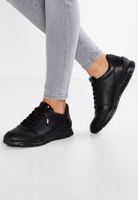 Scarpe sportive in pelle nera con lacci, suola ammortizzata e dettagli di cucitura sottile, abbinate a jeans slim-fit grigi.