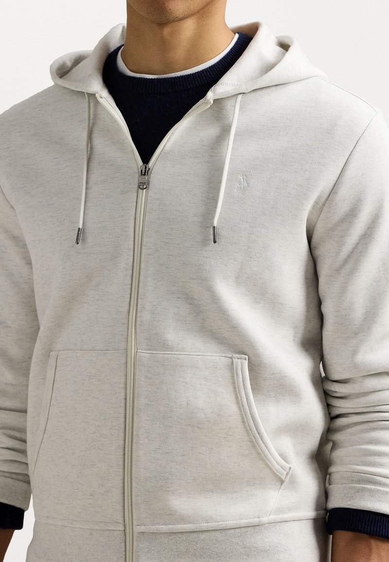 Sudadera con cremallera de color gris claro, con un bolsillo frontal, capucha ajustable y puños acanalados. Presenta un logotipo sutil en el pecho. Material de mezcla de algodón.