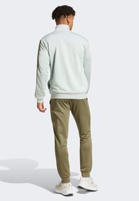 Ljusgrön zip-up jacka med hög krage, kombinerad med olivgröna byxor och vita sportskor. Slät textur och minimalistisk design.