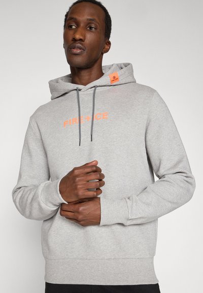 Mann trägt einen hellgrauen Hoodie mit orangefarbenem "FIRE + ICE"-Schriftzug und einem orangefarbenen Label auf der Kapuze, zieht mit beiden Händen den Ärmel zurecht.