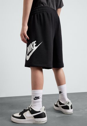 Personne portant un short noir Nike, des chaussettes blanches Nike et des baskets Nike Air noir et blanc debout sur un sol gris avec un fond uni.