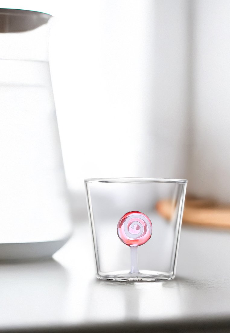 Przezroczysty szklany kubek zawierający małą różowo-białą lizak w kształcie spirali na białej powierzchni z rozmytymi białymi obiektami w tle.