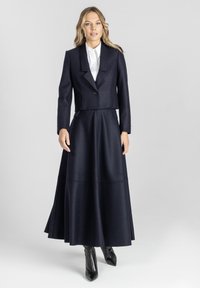 Kobieta w ciemnoniebieskiej dopasowanej marynarce, pasującej szerokiej długiej spódnicy, białej koszuli i czarnych butach na szpilkach na jasnym tle.