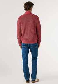 Rød ribbet striktrøje med høj krave, båret med blå denimjeans og beige sko, set fra ryggen.