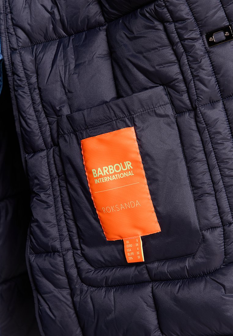 Granatowa kurtka puchowa o pikowanej fakturze. Zawiera pomarańczową metkę z napisem "Barbour International" oraz danymi o rozmiarze. Kieszeń z przodu.