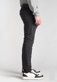 Pantaloni neri slim-fit in denim, con design a cinque tasche, zip laterali e finitura testurizzata. Indossati con sneakers bianche e nere.