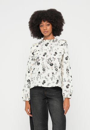 Λευκή μπλούζα με λουλουδένιο μοτίβο, μακριά μανίκια, λεπτομέρειες με βολάν στους ώμους και ψηλό γιακά, σε συνδυασμό με σκούρο τζιν.