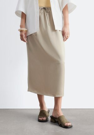 Personne portant une jupe midi beige avec une taille à cordon, une chemise blanche, des sandales à talons blocs ouvertes à l'avant couleur beige, et des bracelets dorés à un poignet.