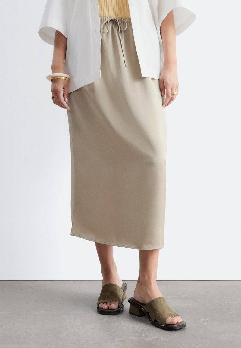Personne portant une jupe midi beige avec une taille à cordon, une chemise blanche, des sandales à talons blocs ouvertes à l'avant couleur beige, et des bracelets dorés à un poignet.