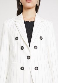 Žena v bílém pruhovaném dvojřadém blejzru se šesti černými knoflíky přes černý žebrovaný top a zlatými náušnicemi.