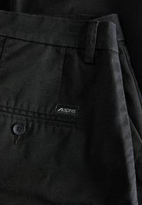 Gros plan d'une poche de pantalon noire avec un bouton et une petite étiquette en tissu indiquant le nom de la marque "apret" sur la couture.