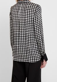 Blusa de patrón de espiga en blanco y negro con un cuello alto, mangas largas y detalles en negro en los puños. Tejido suave y ligero.