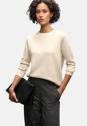 Mujer con cabello rizado, vistiendo un suéter de crema y pantalones de cuadros, sosteniendo un bolso negro en una mano y con la otra mano en el bolsillo, sobre un fondo blanco.