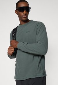 Dunkelgrüner Fleece-Pullover mit rundem Ausschnitt und seitlichen Reißverschlüssen; ausgestattet mit einem gestickten Logo auf der Brust und langen Ärmeln.