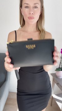 Musta keinonahkainen clutch-laukku, jossa on kultainen varustus, kohokuvioitu logo, vetoketjukiinnitys ja irrotettava rannetuki. Sileä pinta ja minimalistinen muotoilu.