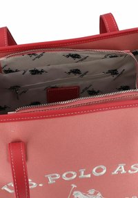 Röd texturerad tygväska med dubbla handtag och dragkedjestängning. Insidan är fodrad med ett ljust tyg som visar ett upprepat logotryck.