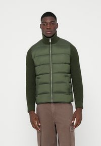 Roheline pehme vesti, millel on kõrge krae ja lukuga sulgur, tekstuursete kootud varrukatega. Seljas on see pruunide cargo-pükste peal, millel on küljetaskud.