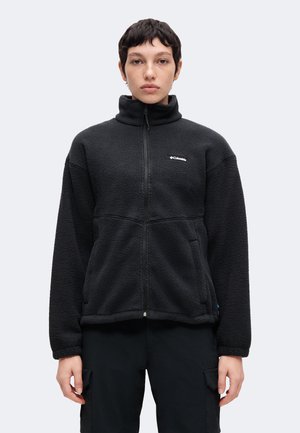 Person mit kurzen dunklen Haaren, die eine schwarze Fleecejacke mit Reißverschluss und schwarze Hose trägt, frontal vor einem einfarbigen Hintergrund steht.
