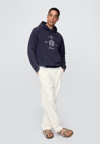 Tengerészeti polár pulóver elején zsebbel és grafikával, krémszínű, bő szabású nadrággal és világosbarna szandállal, pántokkal és csatokkal párosítva.