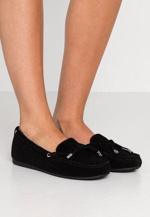 Pieds chaussés de mocassins noirs en daim avec un nœud et des embouts en métal argenté, debout sur une surface lisse gris clair.