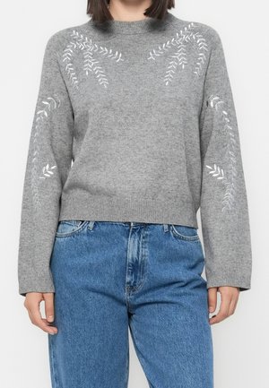 Personne portant un pull en maille gris avec une broderie de feuilles blanches et un jean bleu taille haute, debout devant un fond uni.