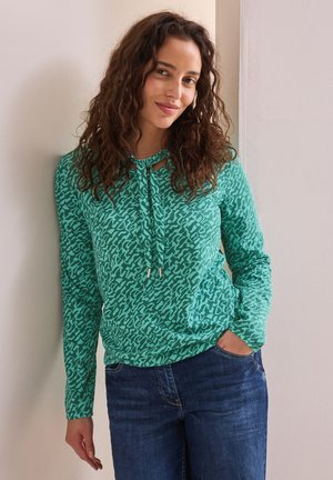 Haut vert à manches longues avec un motif texturé, présentant un décolleté en forme de clé avec des liens, associé à un jean en denim bleu.